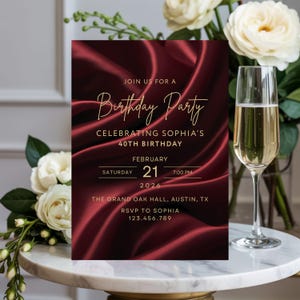 Luxury burgundy silk birthday invitation with gold lettering styled beside champagne glass and flowers