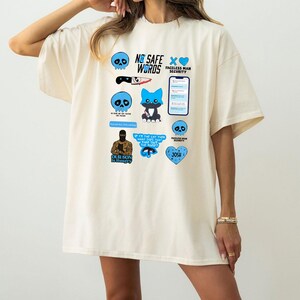 Puede incluir: Camiseta de gran tama&ntilde;o de color crema con varios dise&ntilde;os gr&aacute;ficos azules y negros. Los gr&aacute;ficos incluyen calaveras, un cuchillo, un zorro y texto que dice "NO SAFE WORDS", "FACELESS MAN SECURITY" y "OUR SON IS HUNGRY".