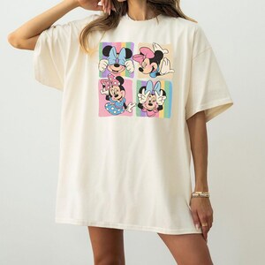 May include: Cream-colored oversized t-shirt featuring a colorful graphic of Minnie Mouse in four different poses. Each pose is set against a pastel rainbow background. The shirt has short sleeves and a relaxed fit.