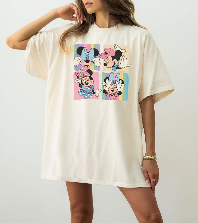 May include: Cream-colored oversized t-shirt featuring a colorful graphic of Minnie Mouse in four different poses. Each pose is set against a pastel rainbow background. The shirt has short sleeves and a relaxed fit.