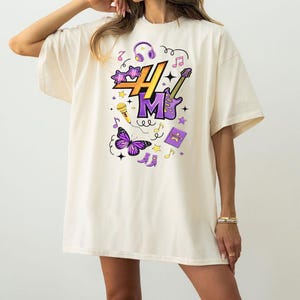 May include: Cream-colored oversized t-shirt featuring a colorful graphic design. The design includes the letters "H" and "M" in purple, along with musical notes, a microphone, a butterfly, and a guitar. The shirt is a casual, comfortable style.
