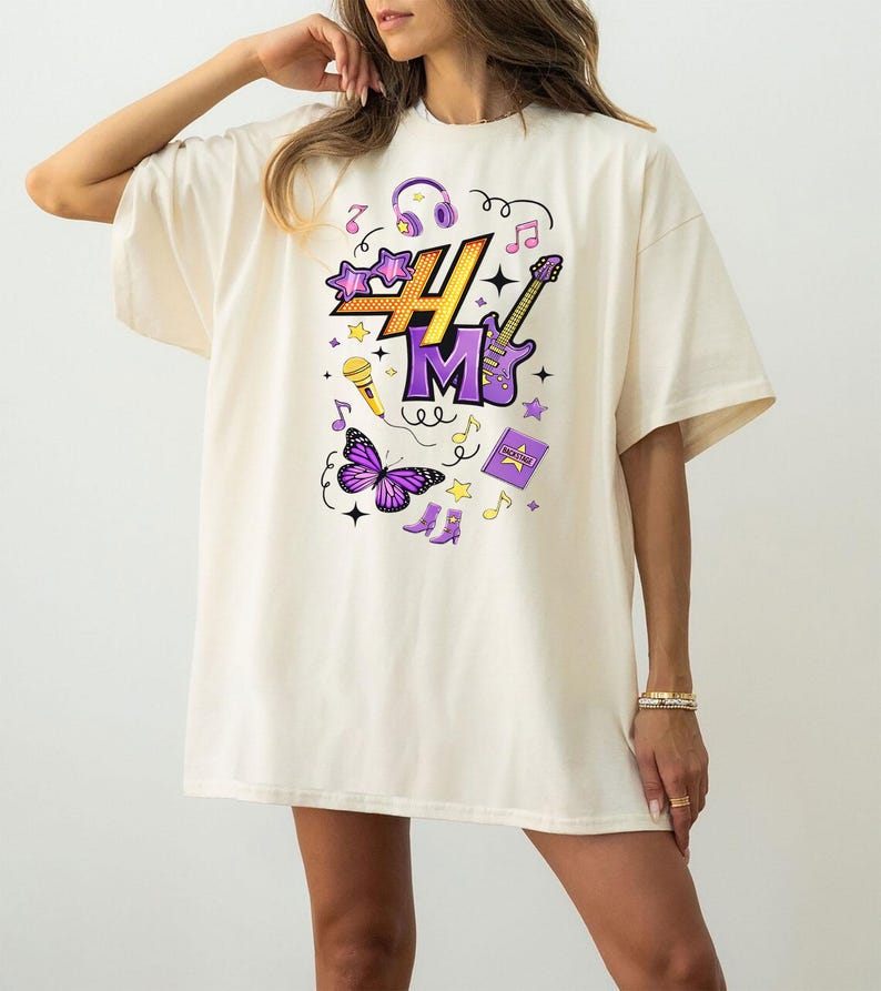 May include: Cream-colored oversized t-shirt featuring a colorful graphic design. The design includes the letters "H" and "M" in purple, along with musical notes, a microphone, a butterfly, and a guitar. The shirt is a casual, comfortable style.