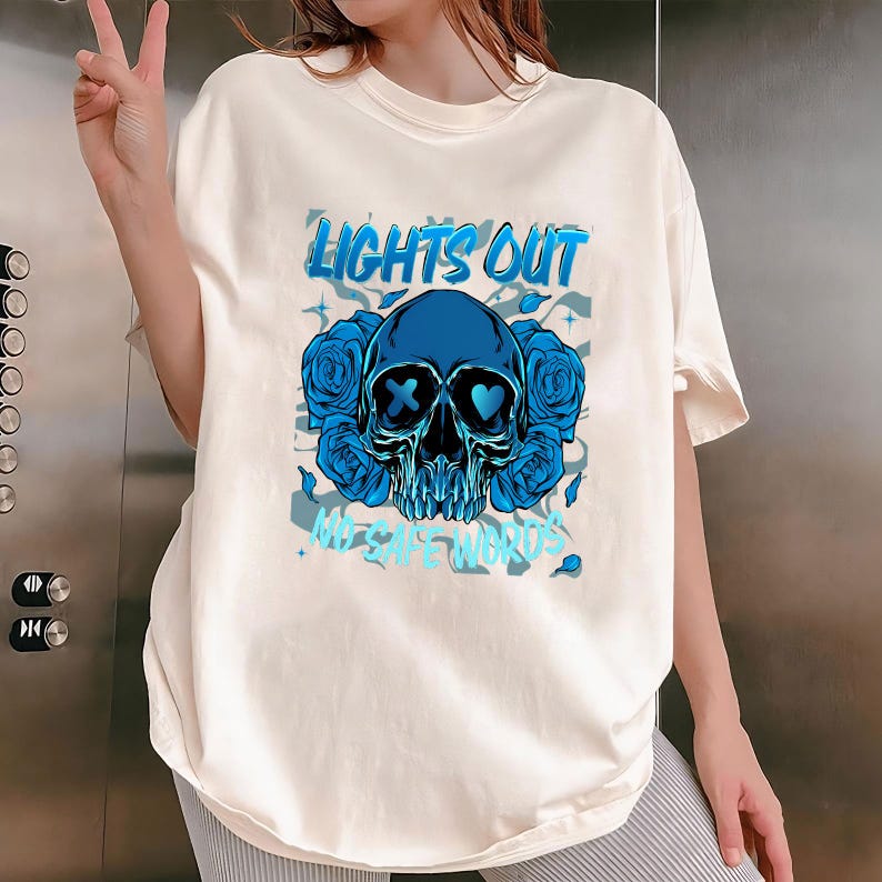 May include: Cream-colored t-shirt featuring a blue skull design with roses and the text "LIGHTS OUT" and "MY SAFE WORDS". The skull has an X and a heart for eyes. The design is surrounded by blue roses.