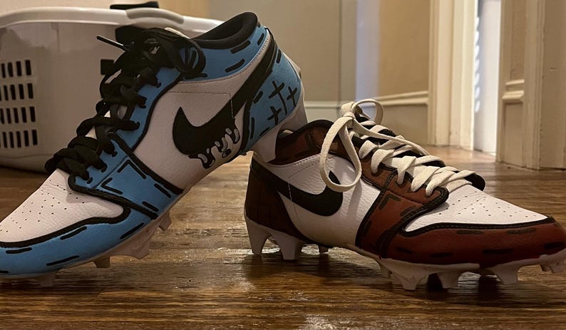 Puede incluir: Par de botas de f&uacute;tbol personalizadas con un dise&ntilde;o de dibujos animados. Una bota presenta una combinaci&oacute;n de colores blanco, azul claro y negro, mientras que la otra es blanca, marr&oacute;n y negra. Ambas tienen cordones negros y un logotipo de Nike.