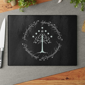May include: Black rectangular cutting board featuring a white tree design with stars and text in a circular pattern. The design is reminiscent of the Lord of the Rings. A knife is visible on the left side of the image.