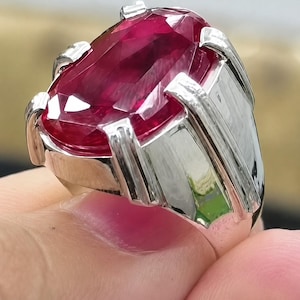 May include: A ring featuring a large, oval-cut, deep red gemstone set in a polished silver-toned metal band. The stone has multiple facets that reflect light, and the setting includes four prominent prongs securing the gem to the band.