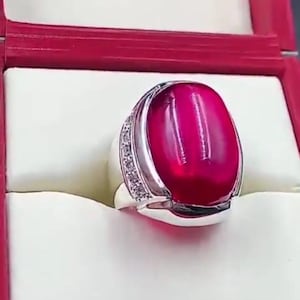 May include: A silver ring with a large, oval, deep red gemstone. The ring has a band of small, clear stones on one side. The ring is presented in a red and white jewelry box.