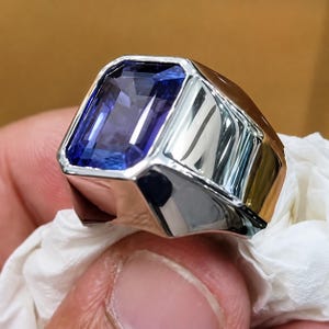 May include: A silver-toned ring featuring a large, rectangular, faceted blue gemstone set in a polished metal band. The stone displays deep violet and blue hues with clear internal reflections, while the band has a smooth, reflective surface.