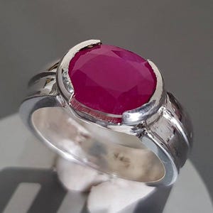 May include: A silver ring featuring a large, faceted, oval-shaped ruby gemstone. The ruby is a deep, vibrant red color. The ring has a simple, yet elegant design with a polished finish.