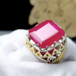 May include: A silver ring with a large, rectangular, ruby-red gemstone. The ring has a detailed design with gold accents and intricate patterns around the band. The gemstone is set in a silver bezel.