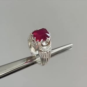 May include: A silver ring with an ornate design and a large, rectangular, deep red gemstone. The ring is held by a silver tool against a neutral background. The gemstone has a faceted surface.