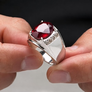 Könnte beinhalten: Ein silberner Ring mit einem großen, oval geschliffenen, tiefroten Edelstein in der Mitte. Die Ringschiene ist aus poliertem Silber und enthält eine Reihe von vier kleinen, klaren, runden Akzentsteinen, die seitlich in die Fassung eingelassen sind.