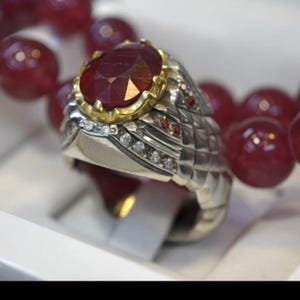 May include: A silver ring with a large, faceted, deep red gemstone set in a gold bezel. The ring is adorned with small diamonds and intricate metalwork. A strand of red beads is in the background.