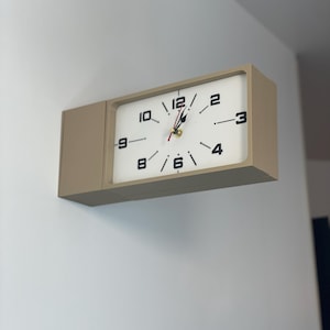 Peut inclure: Une horloge murale rectangulaire avec un cadre beige et un cadran blanc. L'horloge présente des chiffres noirs pour 12, 10, 9, 8, 6, 4, 3 et 2, avec des aiguilles noires pour les heures et les minutes et une trotteuse rouge.