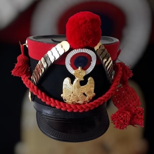Puede incluir: Un sombrero militar negro y rojo con detalles dorados, un pompón rojo y un emblema de águila dorada. El sombrero presenta un cordón trenzado y borlas rojas. Tiene una visera negra y un emblema dorado con una corona.