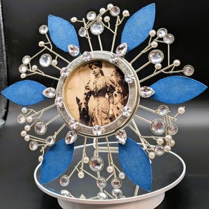 May include: A decorative silver-toned photo frame with a vintage photograph of a woman and dogs. The frame is adorned with blue fabric leaves, clear crystals, and small white pearls. The frame sits on a white, mirrored base.