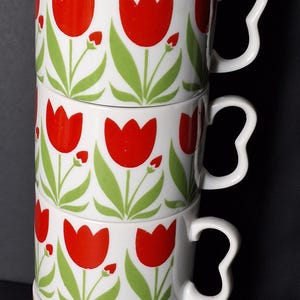 May include: A stack of five white ceramic mugs with a repeating pattern of red tulips and green leaves. Each mug has a white handle. The mugs are stacked vertically against a dark background.