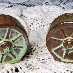 May include: A pair of ceramic salt and pepper shakers. Each shaker is wheel-shaped with a brown top and a light green and brown wheel design. The shakers are on a white, lacy surface.