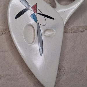 May include: A cream-colored ceramic wall mask with a speckled finish and an elongated, asymmetrical shape. The surface features a graphic design with black lines, a red triangle, and a blue oval. It includes two oval cutouts and a pointed base.