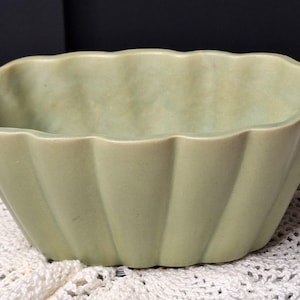 May include: A light green ceramic planter with a scalloped rim and vertical ribbed sides. The planter has a rounded rectangular shape and sits on a white lace surface. The color is a muted, vintage green.