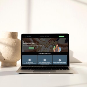 May include: A laptop displays a website with the text "Become the person you were meant to be." The website offers life vision, career achievement, and purpose-oriented coaching. A beige vase sits to the left of the laptop.