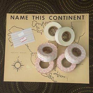 May include: A beige educational map titled NAME THIS CONTINENT with a compass rose, featuring five rolls of adhesive tape. Two rolls are solid white, and three rolls feature a repeating red pattern. A small rectangular index card with a red SALE sticker.