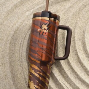 Puede incluir: Vaso Stanley de acero inoxidable con asa y pajita. Presenta un dise&ntilde;o de rayas de tigre marr&oacute;n y dorado. El logotipo de Stanley es visible en el lateral. Descansa sobre una superficie arenosa.