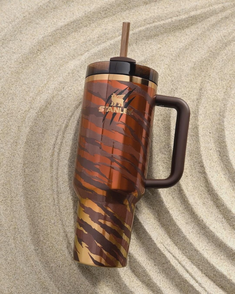 Puede incluir: Vaso Stanley de acero inoxidable con asa y pajita. Presenta un dise&ntilde;o de rayas de tigre marr&oacute;n y dorado. El logotipo de Stanley es visible en el lateral. Descansa sobre una superficie arenosa.