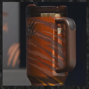 Puede incluir: Taza de viaje de acero inoxidable con un estampado de rayas de tigre en tonos naranja y marr&oacute;n. La taza tiene un asa, tapa y ribete marr&oacute;n oscuro. El logotipo de Stanley es visible.