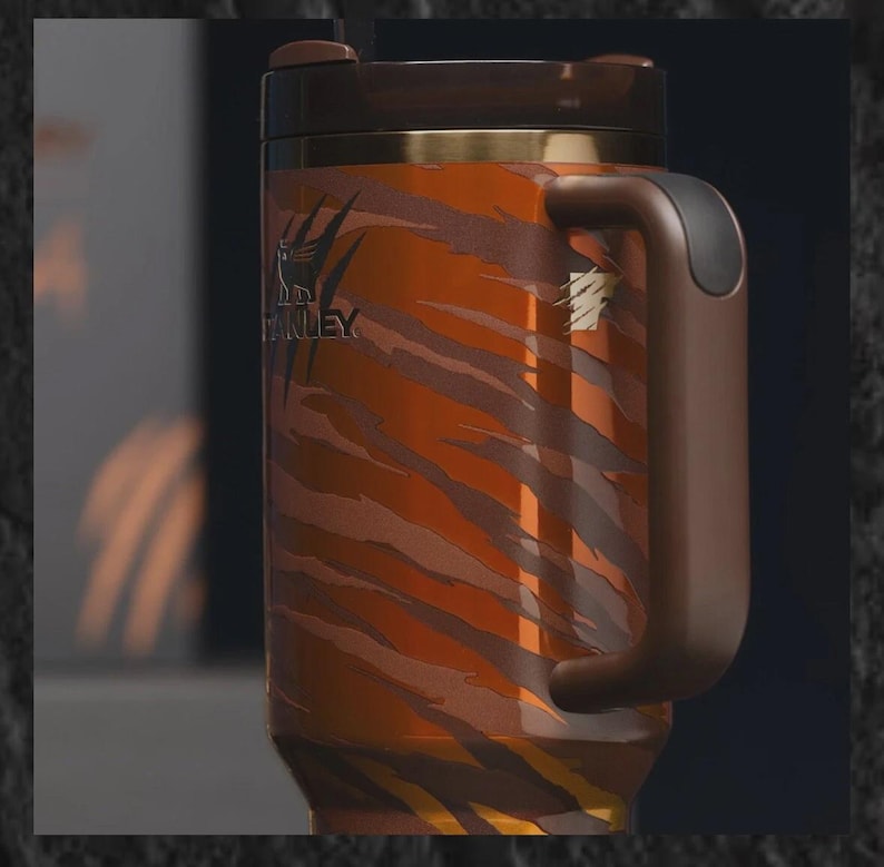 Puede incluir: Taza de viaje de acero inoxidable con un estampado de rayas de tigre en tonos naranja y marr&oacute;n. La taza tiene un asa, tapa y ribete marr&oacute;n oscuro. El logotipo de Stanley es visible.
