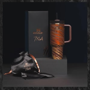 Puede incluir: Un vaso Stanley TYLA con un dise&ntilde;o de rayas de tigre en tonos marr&oacute;n y dorado, una tapa y pajita negras y un asa de oro rosa. El vaso se exhibe con una caja de regalo negra y una bolsa de sat&eacute;n negra, ambas con los logotipos de Stanley y TYLA.