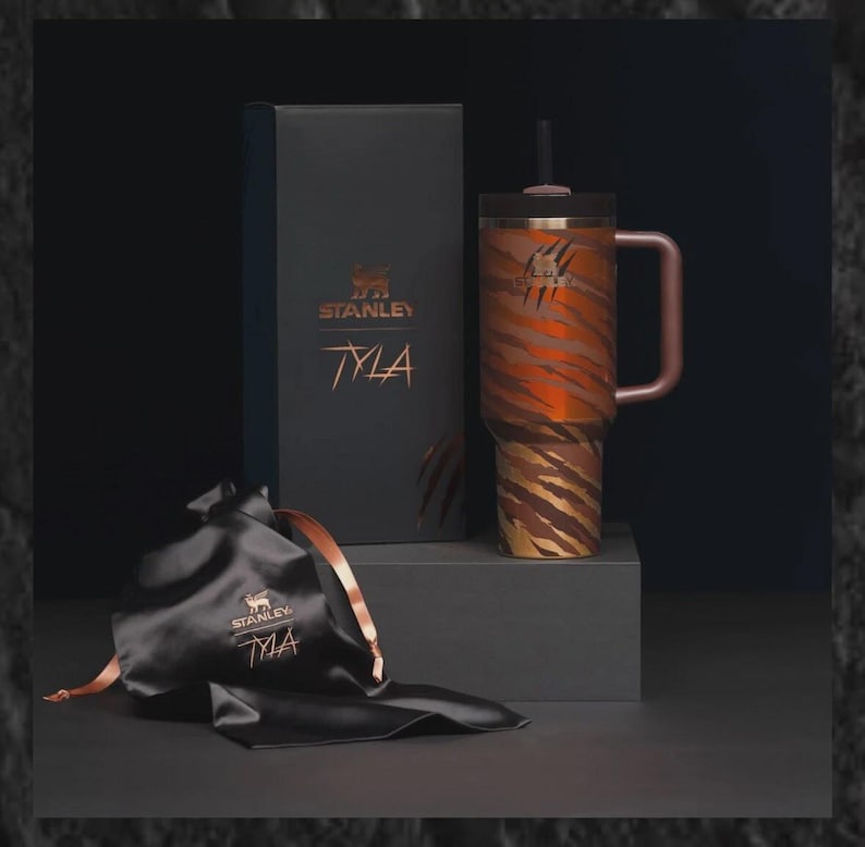 Puede incluir: Un vaso Stanley TYLA con un dise&ntilde;o de rayas de tigre en tonos marr&oacute;n y dorado, una tapa y pajita negras y un asa de oro rosa. El vaso se exhibe con una caja de regalo negra y una bolsa de sat&eacute;n negra, ambas con los logotipos de Stanley y TYLA.