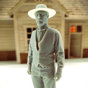 May include: A gray-colored miniature figurine of a man wearing a hat, vest, long-sleeve shirt, and pants. The figure is standing in front of a miniature building. The hat has a wide brim.