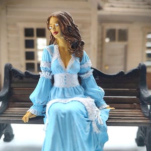 May include: A figurine of a woman in a long, light blue dress with white lace trim, seated on a black bench. The dress features long sleeves and a low neckline. The woman has brown hair and is looking to the side.