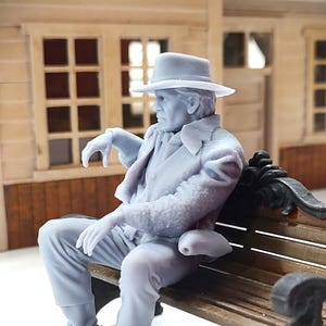 May include: A light gray figurine of a person seated on a wooden bench. The figure wears a hat, coat, gloves, and boots. The person is posed with one arm resting on the bench and the other arm resting on their lap. The background includes a building with windows.
