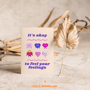 May include: A greeting card with the text "It's okay to feel your feelings" and illustrations of cartoon hearts. The hearts are in various colors and expressions. The card is beige with purple and pink text.