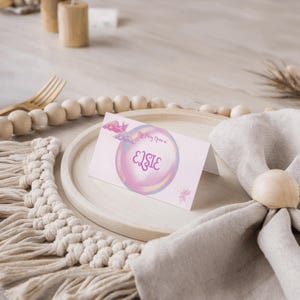 Könnte beinhalten: Eine hellrosa Tischkarte mit dem Namen "Elsie" in einem Blasendesign. Die Karte enthält auch den Text "My Fairy Name is:" und eine kleine Schmetterlingsillustration. Die Karte liegt auf einem Holztablett mit einem Makramee-Platzset.