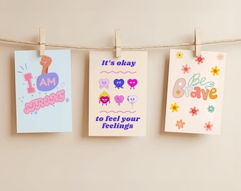 May include: Three art prints hanging from a string with wooden clips. The prints feature positive affirmations and cute illustrations. One says "I AM STRONG", another says "It's okay to feel your feelings", and the last says "Be Brave".