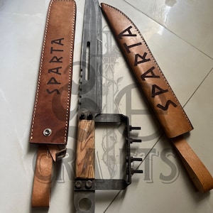 May include: A large, silver knife with a wooden handle and a metal guard, alongside two brown leather sheaths. The word "SPARTA" is embossed on each sheath. The knife blade has a serrated edge.