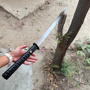 以下が含まれることがあります： 黒と銀色の刀、テクスチャ加工された柄付き。 刃は銀色で、柄は黒に金色のアクセント。 自然な屋外の背景に対して刀が保持されています。