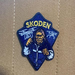 May include: A navy blue patch with the word "SKODEN" in yellow lettering. The patch features an illustration of a person in a blue jacket with their fists clenched. The patch is diamond-shaped with a blue border and white accents.