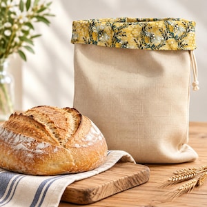 May include: A beige fabric bread bag with a drawstring closure and a folded-over top cuff featuring a yellow and green floral pattern. The bag is positioned next to a round, crusty loaf of bread on a striped cloth and wooden board.
