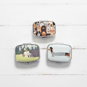 May include: Three rectangular metal pill boxes with hinged lids. One box features a crowd of various dog breeds. Another depicts a sleeping cat. The third shows a dachshund wearing a striped shirt and a beret.