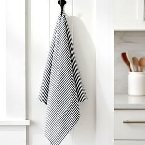 May include: A rectangular kitchen towel featuring a pattern of thin, vertical black and white stripes. The fabric is draped and hangs from a black metal hook mounted on a white paneled surface, displaying a consistent striped textile design.