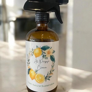 May include: Amber glass spray bottle with a black trigger. The label features a lemon and floral wreath design with the words "All Purpose Cleaner", "Fresh Natural Sustainable". The bottle is designed for cleaning.