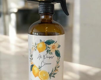May include: Amber glass spray bottle with a black trigger. The label features a lemon and floral wreath design with the words "All Purpose Cleaner", "Fresh Natural Sustainable". The bottle is designed for cleaning.