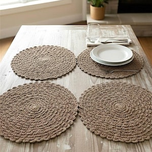 May include: Four round, brown crochet placemats on a light wood table. One placemat has white plates, silverware, and a napkin. A glass and a potted plant are also on the table. The placemats have a textured, circular design.