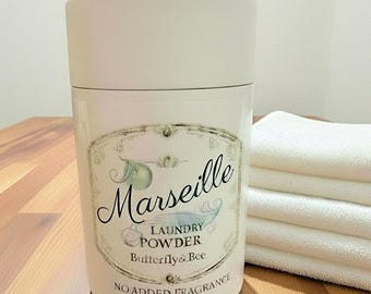 May include: A cylindrical white container with a lid, featuring a label that reads Marseille LAUNDRY POWDER Butterfly & Bee NO ADDED FRAGRANCE. The label includes a decorative border and illustrations of a green fruit and a small flower.