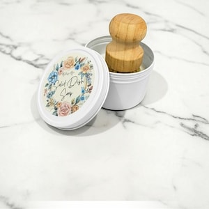 May include: A white metal tin containing solid dish soap with a wooden scrub brush resting inside. The lid features a floral wreath design surrounding the text Solid Dish Soap. The container is open, showing the brush handle and the soap surface.