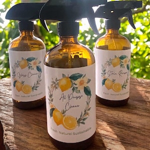 May include: Three amber glass spray bottles with black spray nozzles. Each bottle has a white label with a lemon and floral design. The labels read "Glass Cleaner", "All Purpose Cleaner", and "Stain Remover". The text "Fresh Natural Sustainable" is also on the labels.